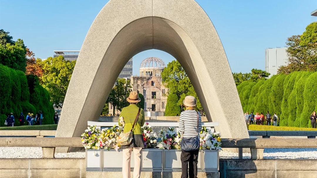 Tourists at Hiroshima Victims Memorial Cenotaph with Atomic Bomb Dome in view.