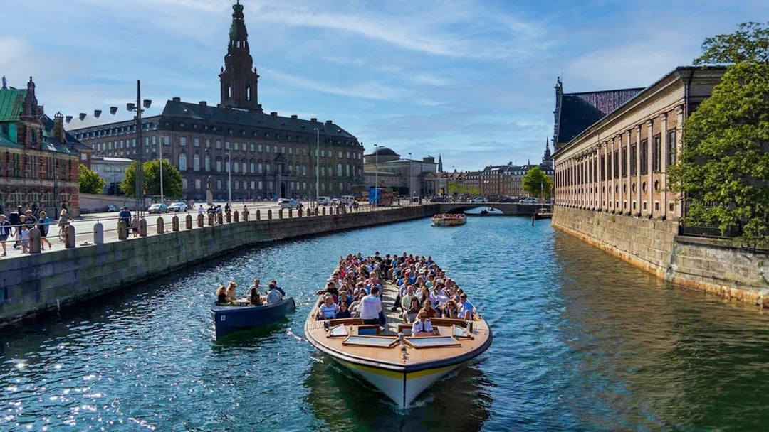 Canal cruise boat with tourists in Copenhagen, passing historic buildings and a bridge.