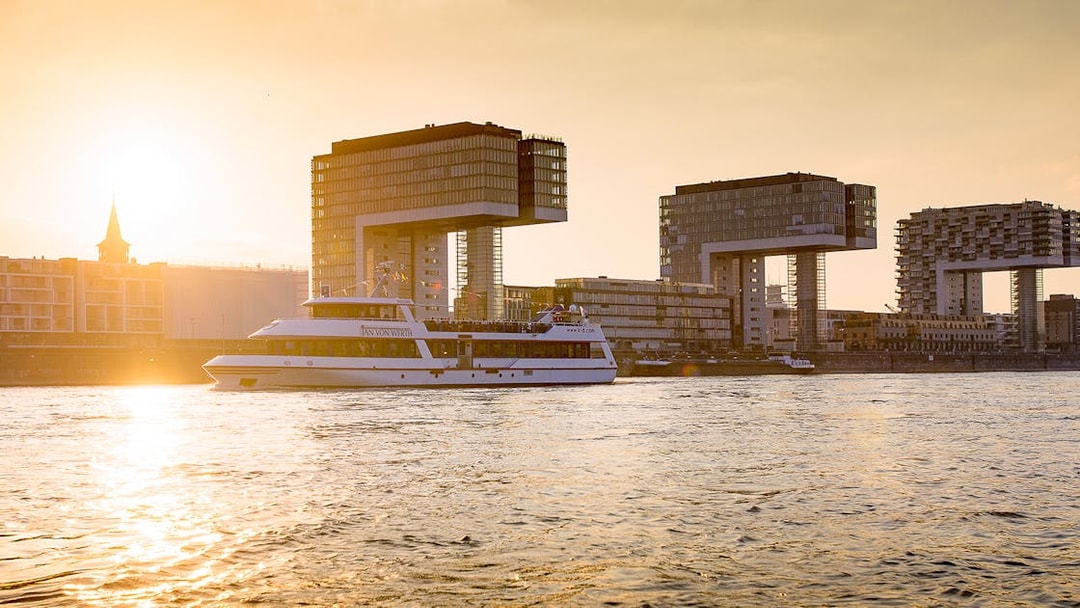 Cruise boat on the Rhine River with Cologne's Kranhäuser buildings at sunset.