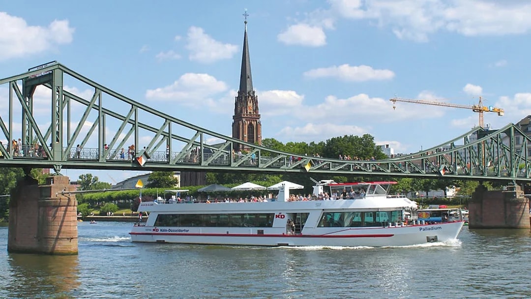 Cruise boat passing under a bridge in Frankfurt with a church spire in the background.