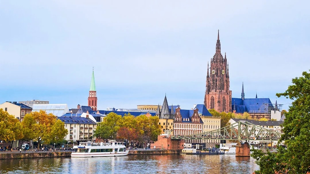 Cityscape of Frankfurt with a cruise boat on the Main River, featuring the Frankfurt Cathedral.