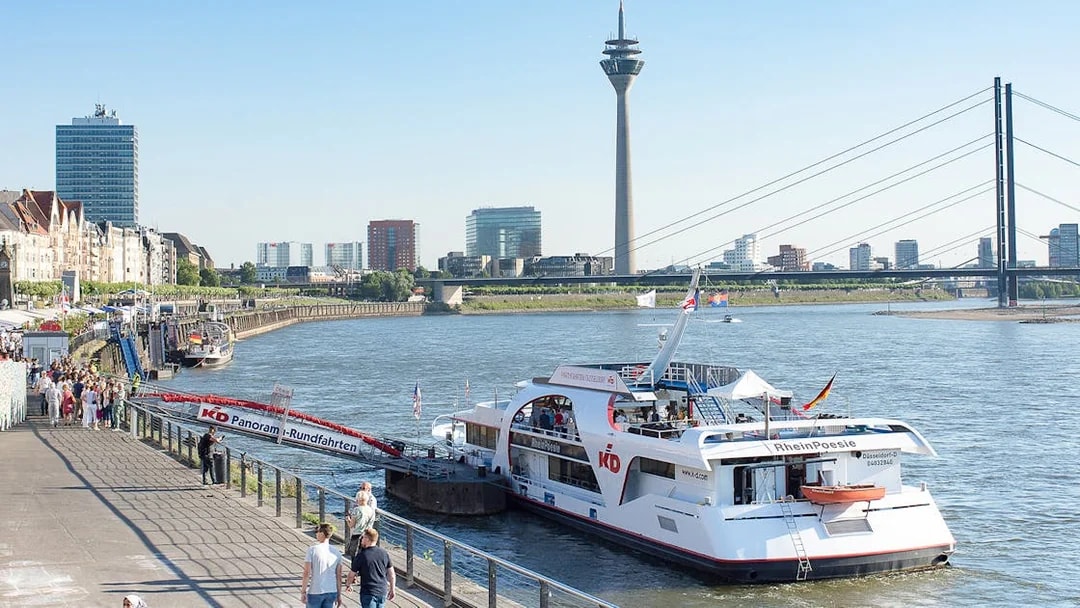 Panorama cruise boat docked on the Rhine River with Düsseldorf skyline and Rheinturm tower.