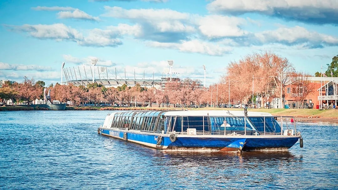 River cruise boat on a scenic waterway with a stadium and trees in the background.