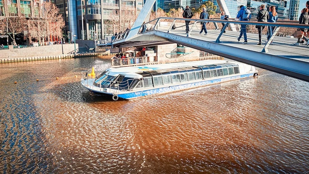 River cruise boat passing under a pedestrian bridge in a cityscape setting.