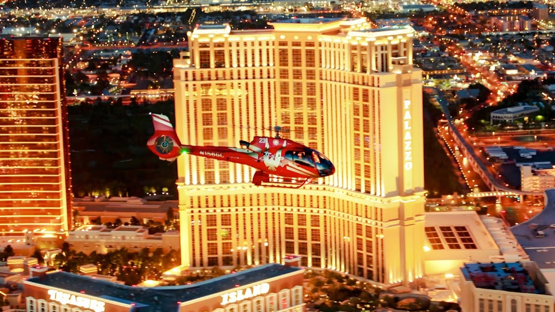 Red helicopter flying over Las Vegas Strip at night with Palazzo Hotel and Treasure Island in view.