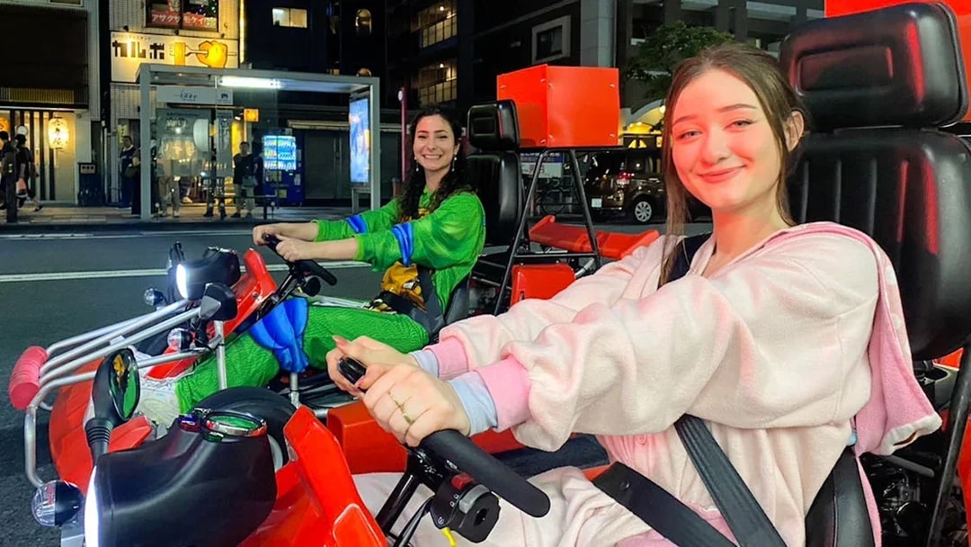 Two people in costumes driving go-karts on a street in Osaka Namba at night.