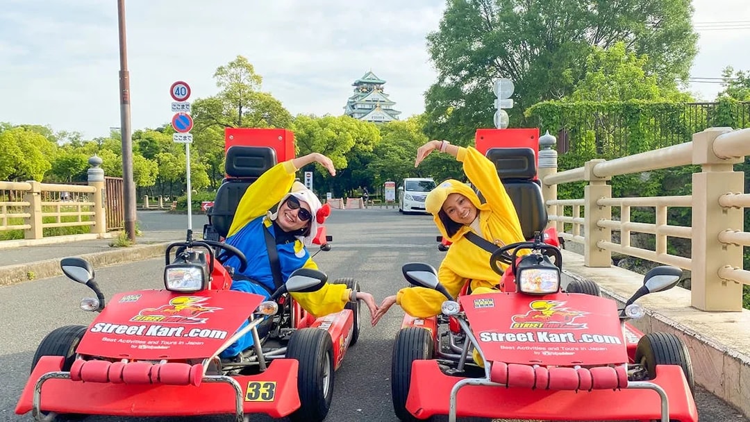 Go-kart riders in costumes pose on a street in Osaka Namba with Osaka Castle in the background.