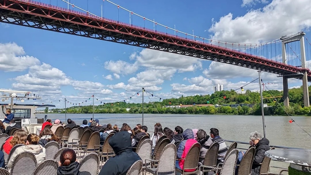 Tourists seated on a boat during a Bordeaux sightseeing river cruise under a large bridge.