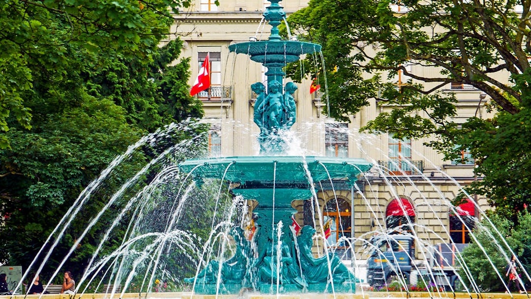 Fountain in Geneva with Swiss flags and historic building in the background.