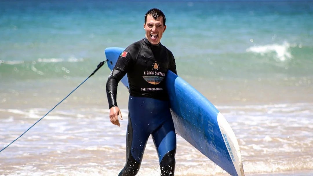 Surfer carrying a blue surfboard on a beach during a group surf lesson in Lisbon.