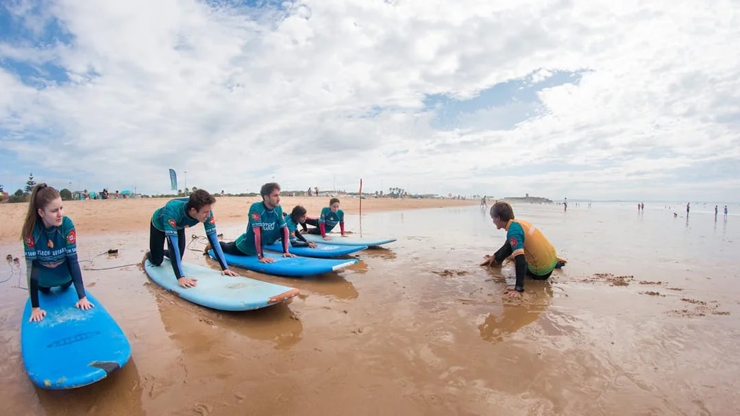 Group surf lesson on a sandy beach with instructor guiding participants on surfboards.