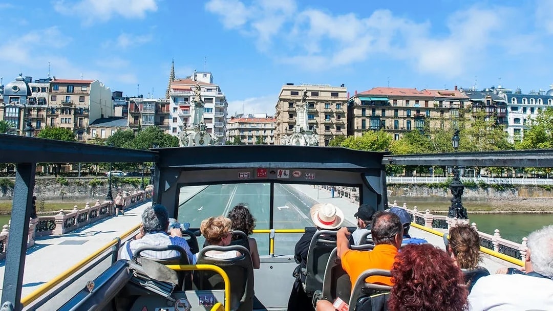 Open-top bus tour in San Sebastian city with passengers viewing historic architecture.