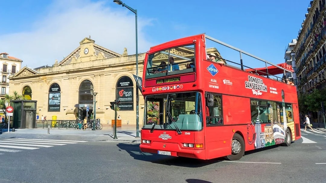 Red double-decker bus on San Sebastian city tour near historic building.