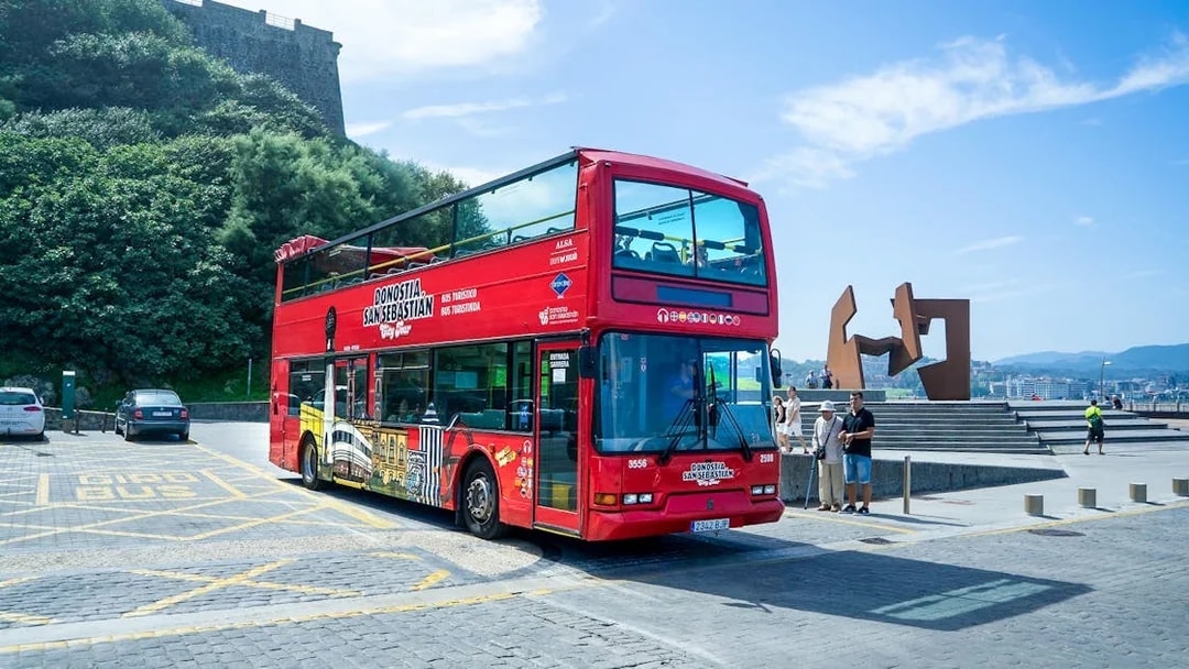 Red double-decker tour bus in San Sebastian near a modern sculpture and scenic view.