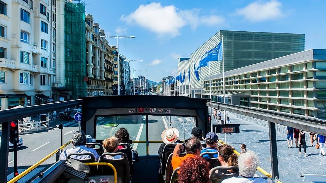 Open-top bus tour in San Sebastian city with passengers viewing modern architecture.