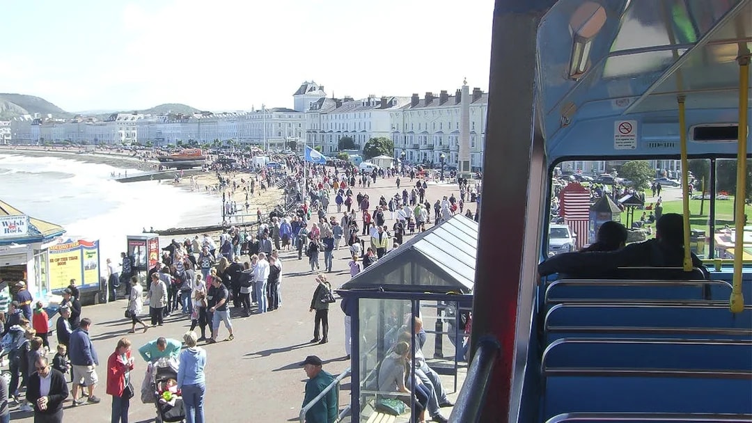 Open-top bus view of Llandudno promenade with crowds and beach, part of 24-Hr Hop-On-Hop-Off Tour.