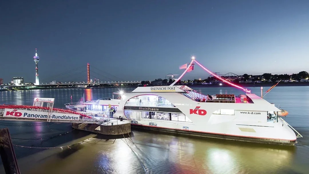 Evening cruise ship on the Rhine River with Düsseldorf skyline and Rheinturm in the background.