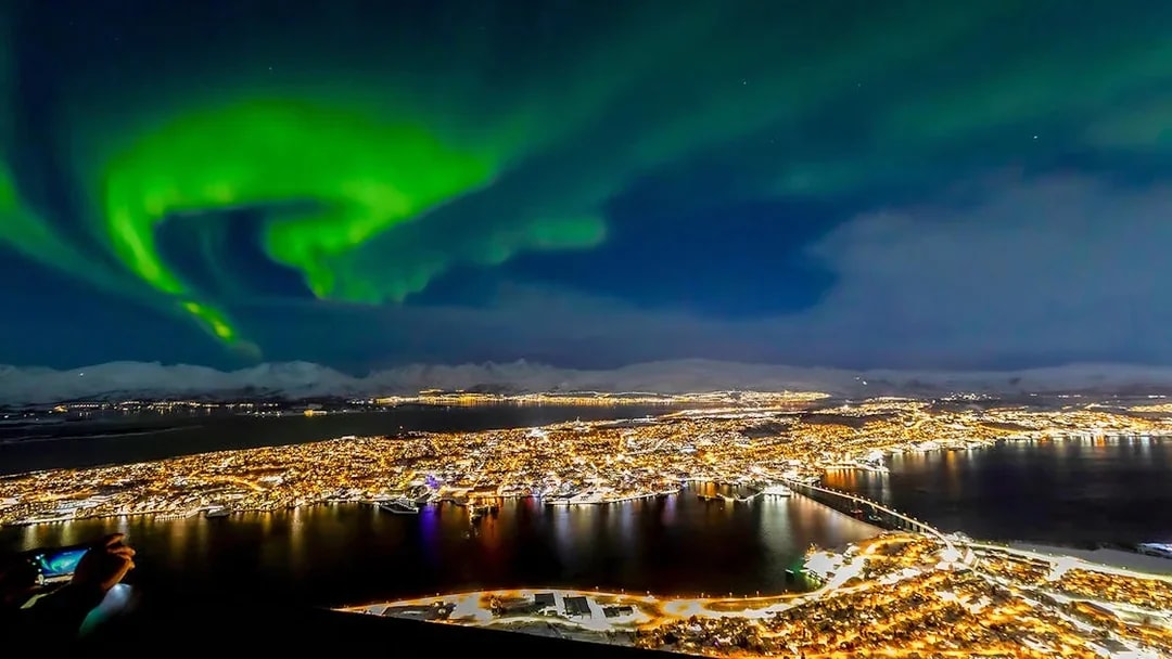 Northern lights over Tromso, Norway, with city lights reflecting on the water.