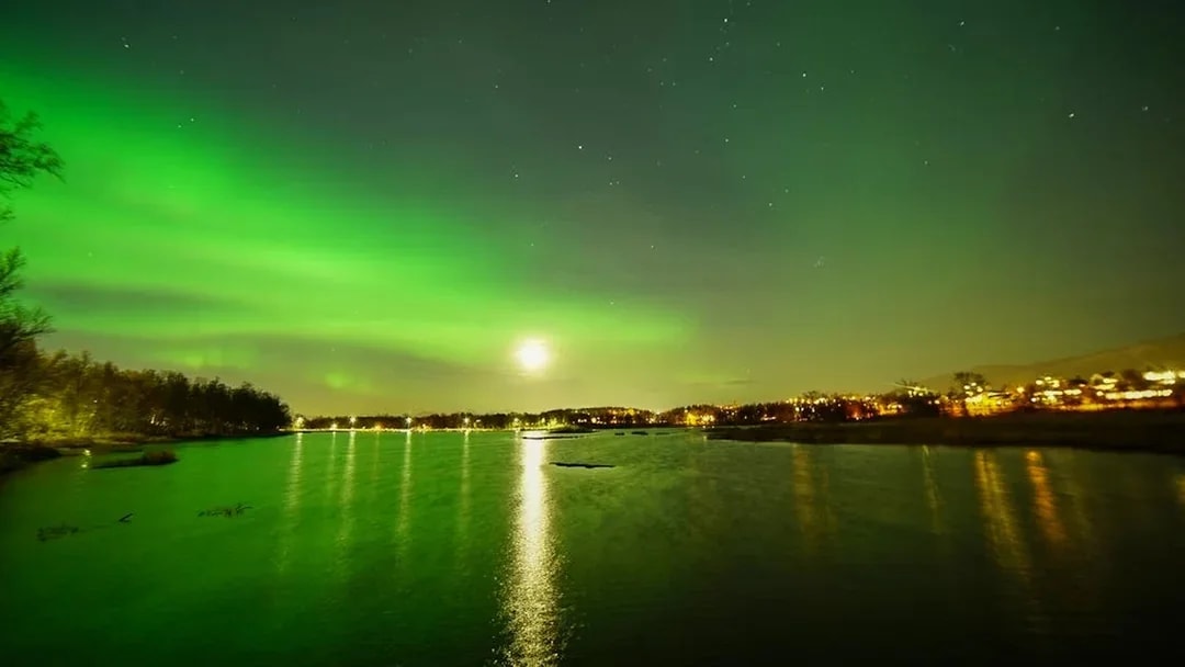 Aurora borealis over a calm lake with distant city lights reflecting on the water.