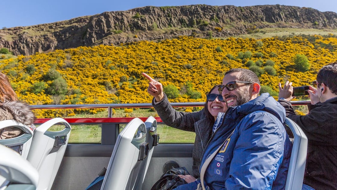 Passengers enjoying the view from the upper deck of a HOHO bus near a hillside with yellow flowers.