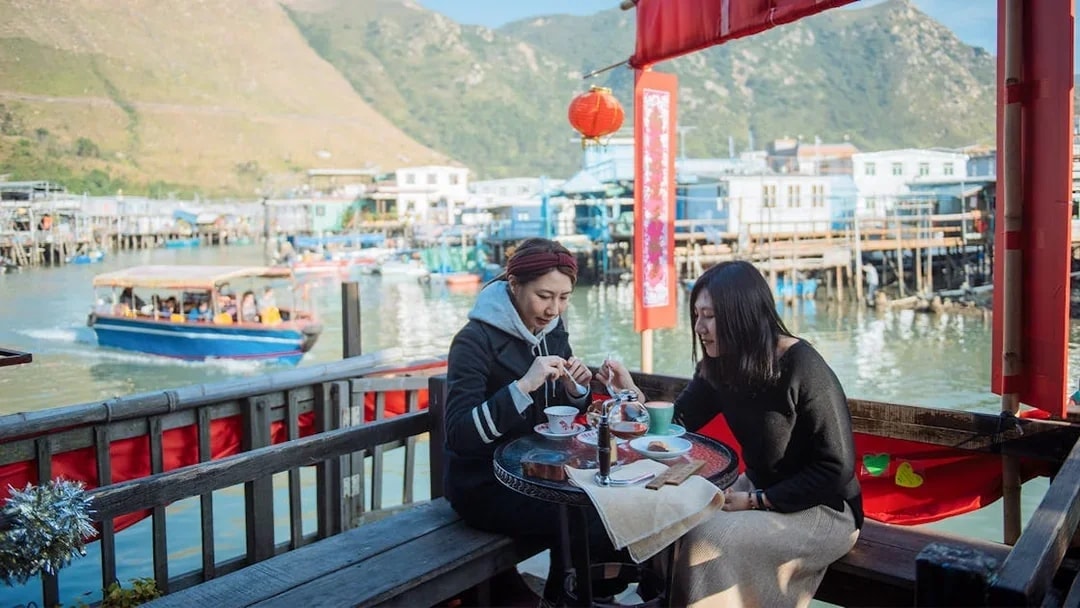 Two people dining at a waterfront café in Tai O, Hong Kong, with stilt houses and boats in the background.