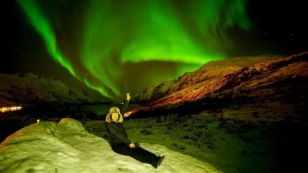 Tourist sitting on snowy hill under Northern Lights in Tromso, Norway.