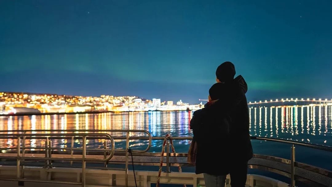 Couple on a cruise ship viewing Northern Lights over a lit cityscape.