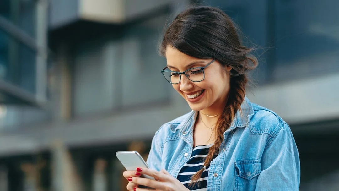 Woman smiling while reading a message on her phone outdoors.