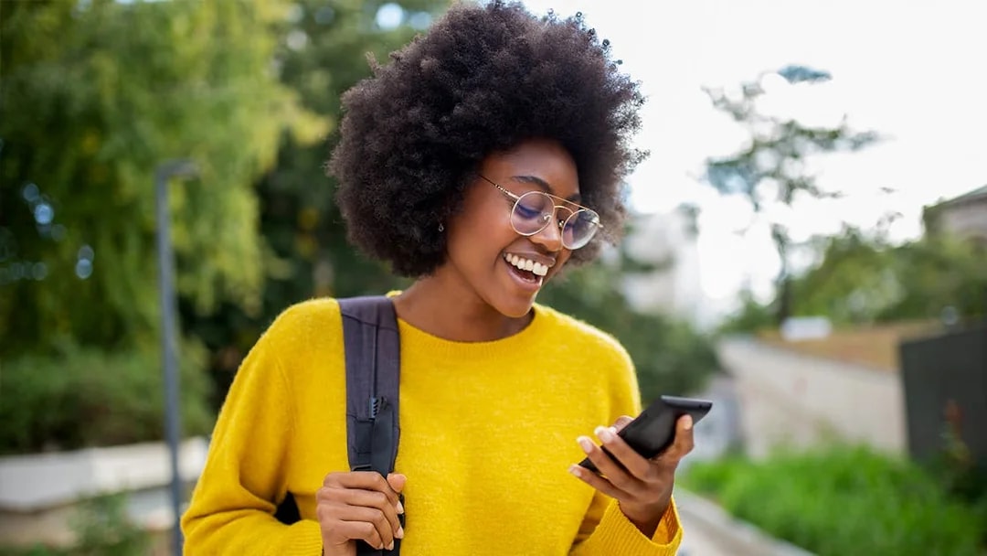 Woman with glasses smiling while checking her phone outdoors.
