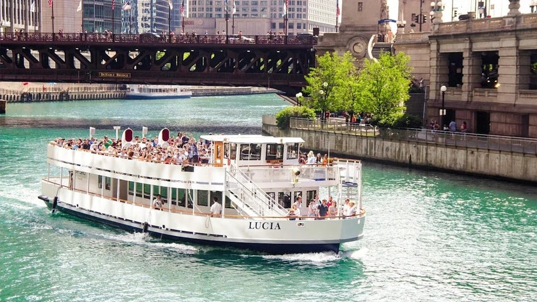 Chicago River cruise boat passing under DuSable Bridge with city buildings in the background.