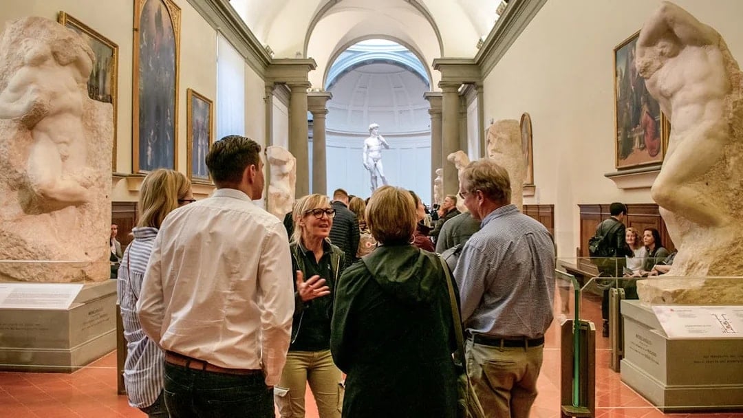 Guide leading tour group in Accademia Gallery, Florence, with Michelangelo's David in background.