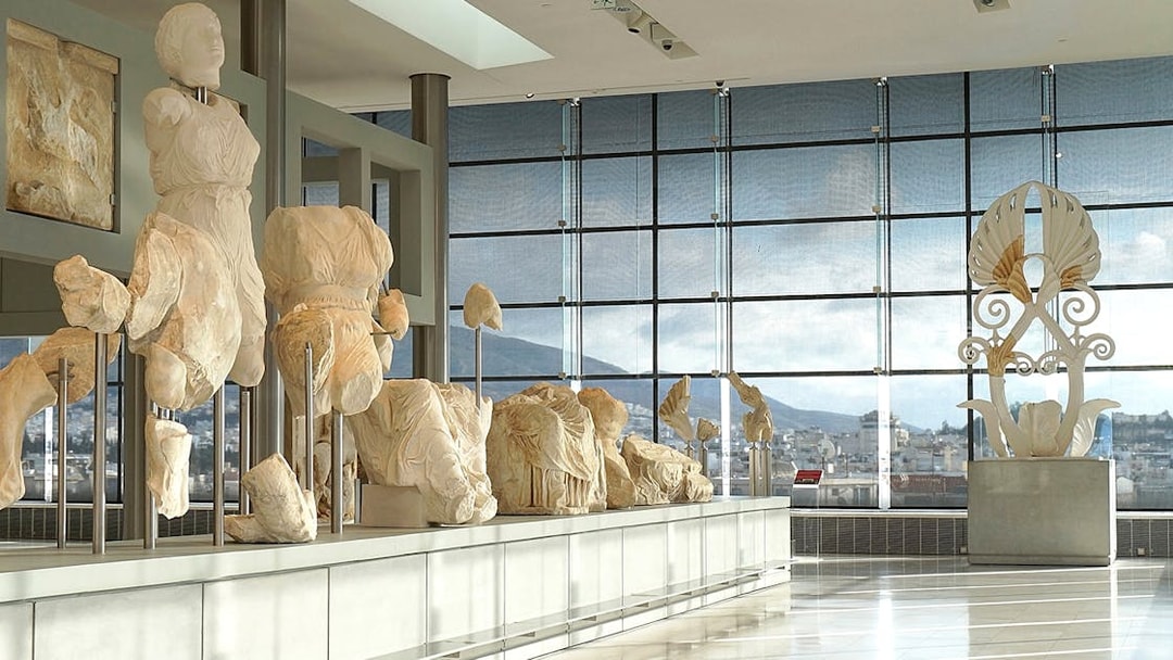 Ancient sculptures displayed in the Acropolis Museum with city view through large windows.