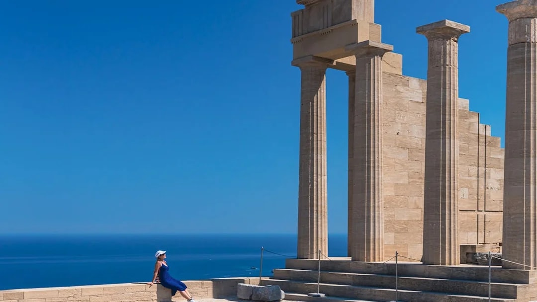 Ancient columns at Acropolis of Lindos with a person sitting nearby, overlooking the sea.