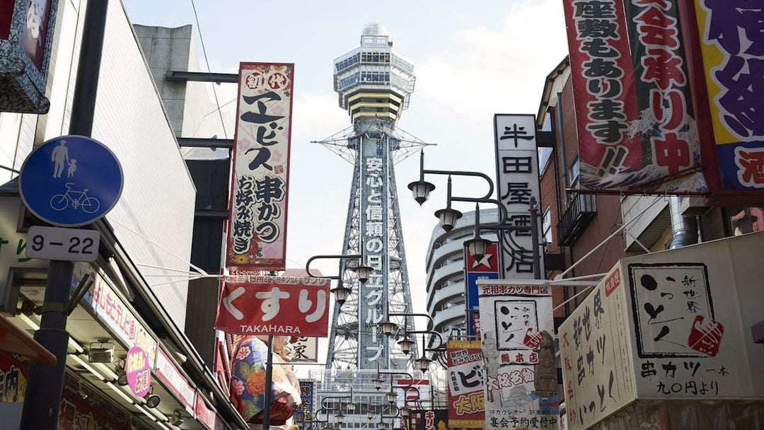 Tsutenkaku Tower in Osaka, Japan, viewed from a bustling street with colorful signs.