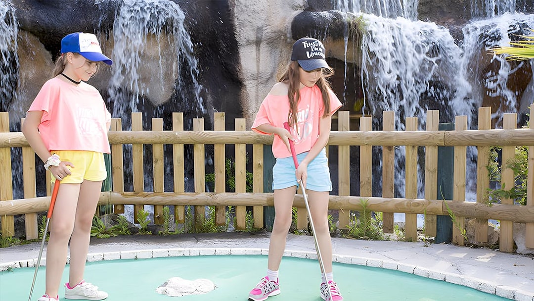 Kids playing mini-golf near a waterfall at an adventure golf course.