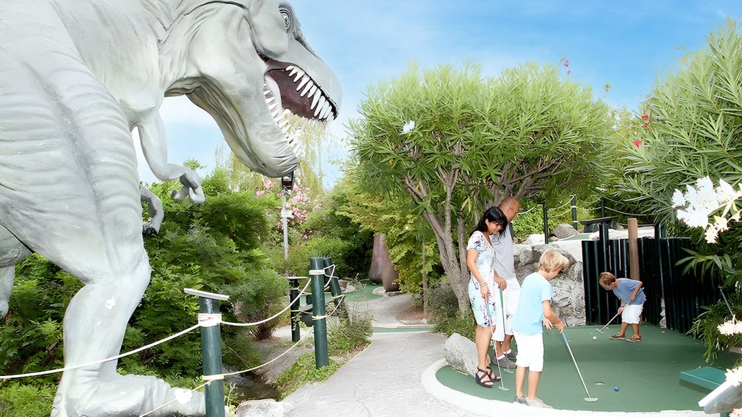 Family playing mini-golf near dinosaur statue at adventure park.