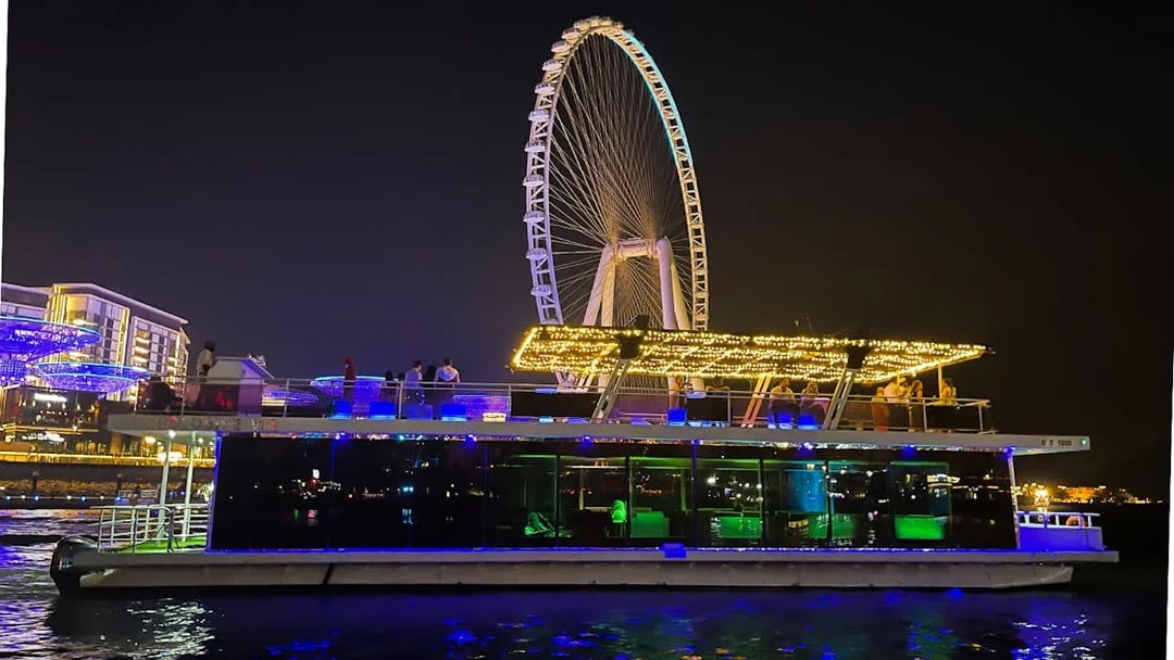 Houseboat with lights on water near Ain Dubai Ferris wheel at night.