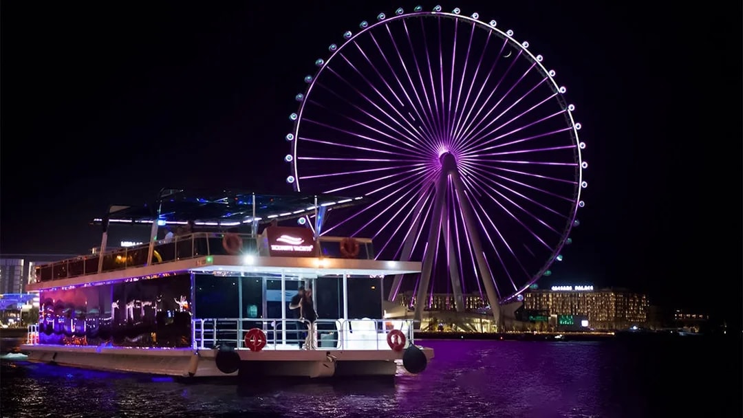 House boat on water with Ain Dubai Ferris wheel illuminated at night.