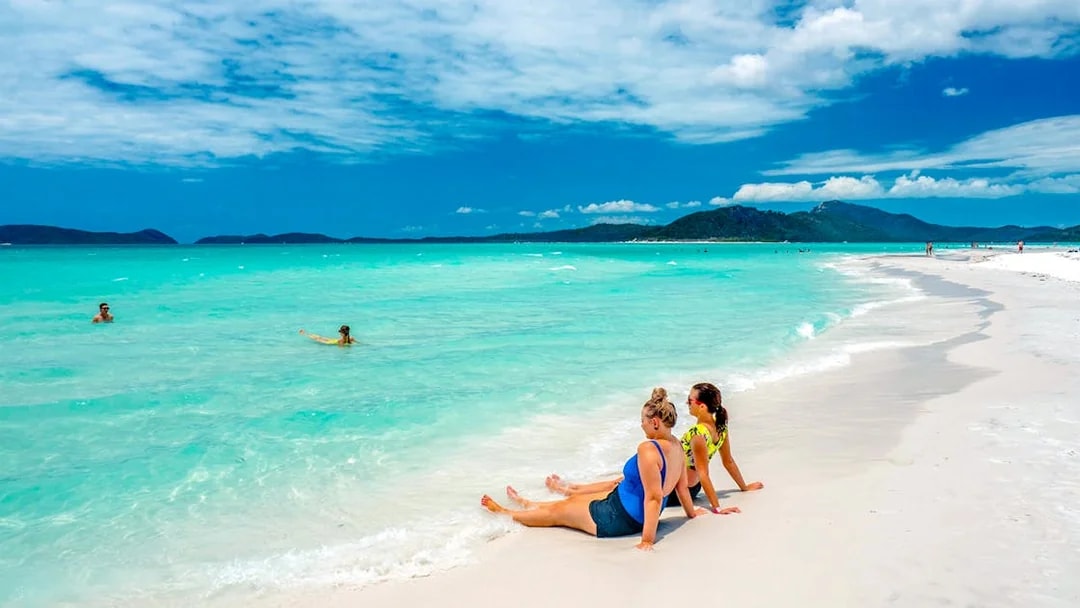 Tourists relaxing on Whitehaven Beach with turquoise water and distant hills.
