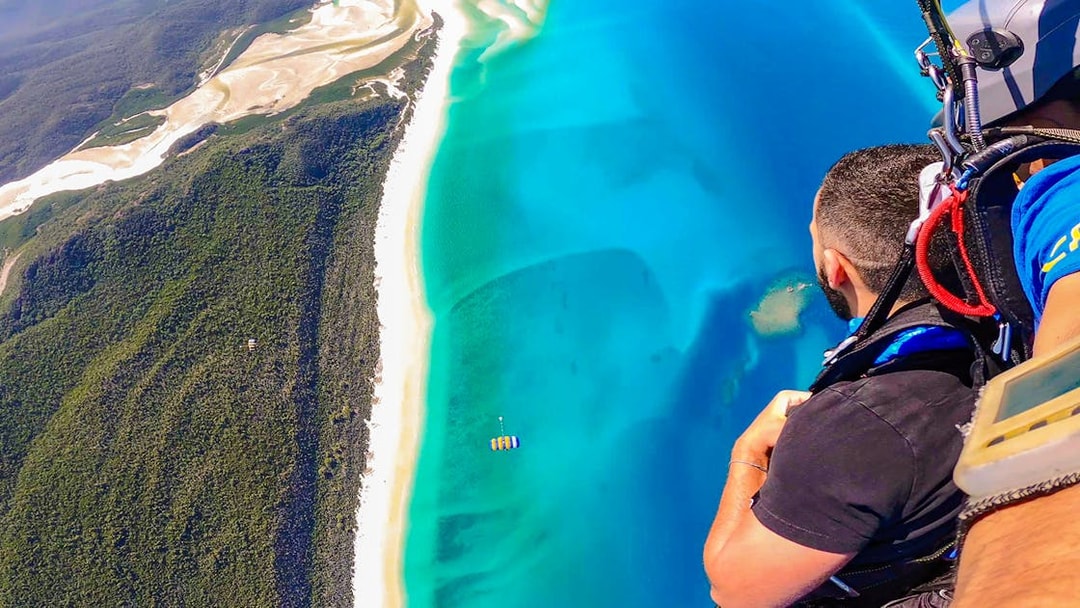 Tandem skydivers over Airlie Beach coastline with ocean and clear skies.