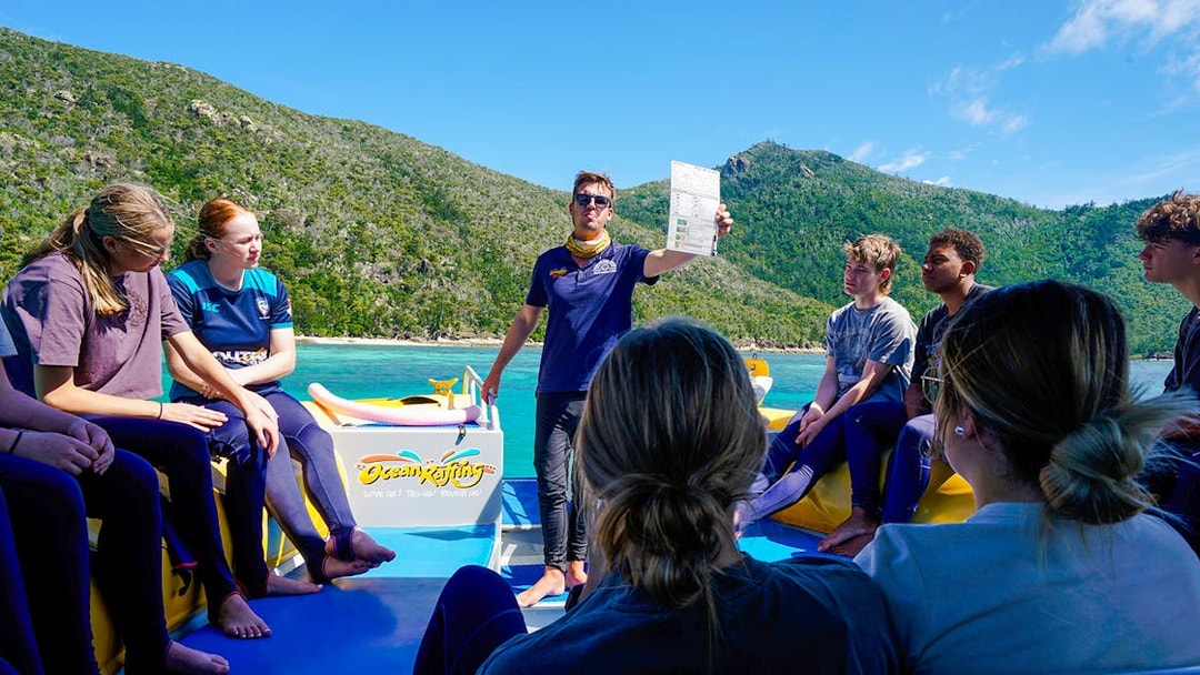 Group on a boat tour near Whitsundays, guide holding a paper, lush hills in the background.