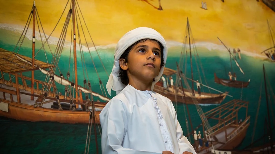 Child in traditional attire at Al Shindagha Museum with dhow mural in background.