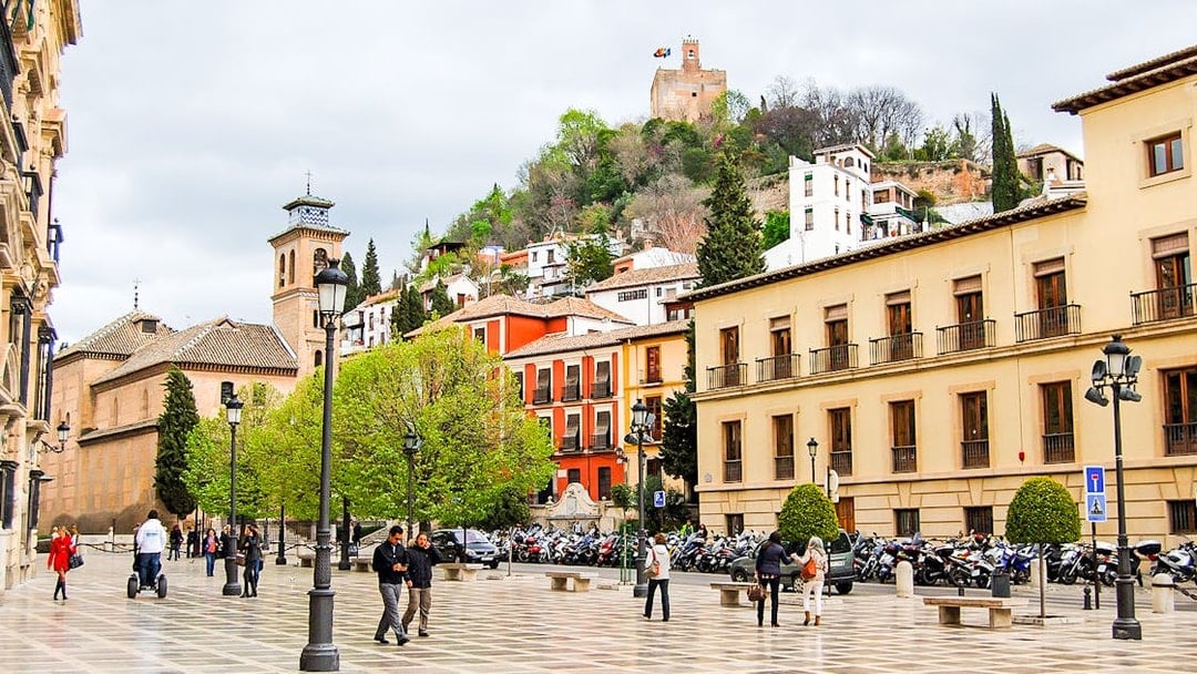 Plaza in Albaicin with view of Sacromonte, Granada, featuring historic buildings and people walking.