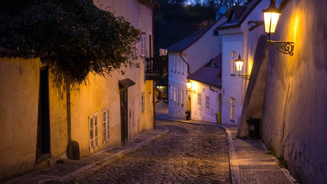 Narrow cobblestone street in Prague Castle area at dusk, illuminated by street lamps.
