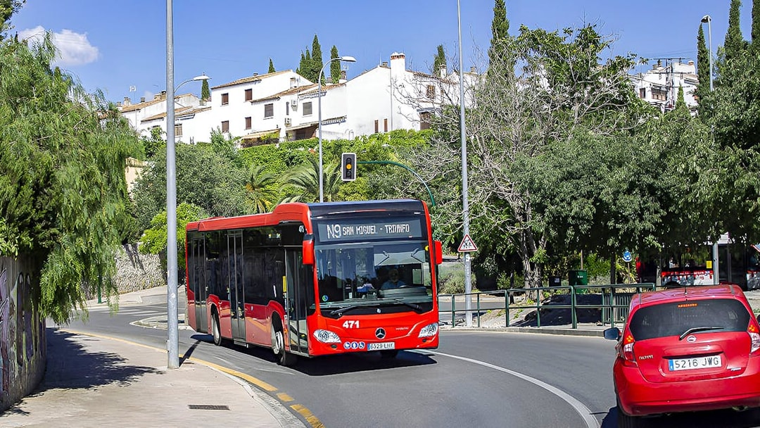 Granada city bus on route near Alsa station, surrounded by greenery and buildings.