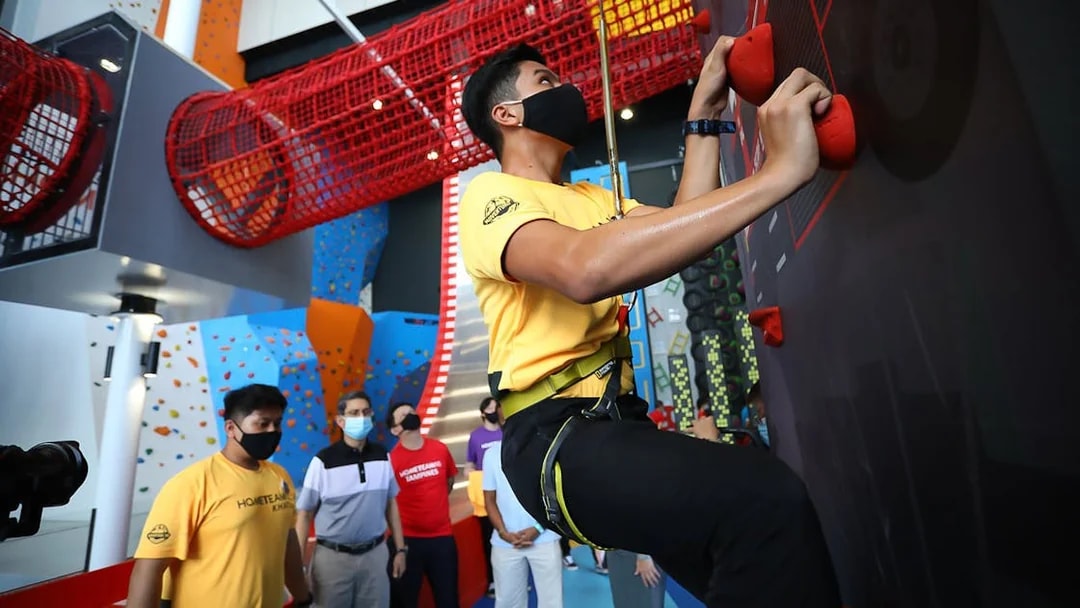 Climber scaling indoor wall at Adventure HQ with red net tunnel above.