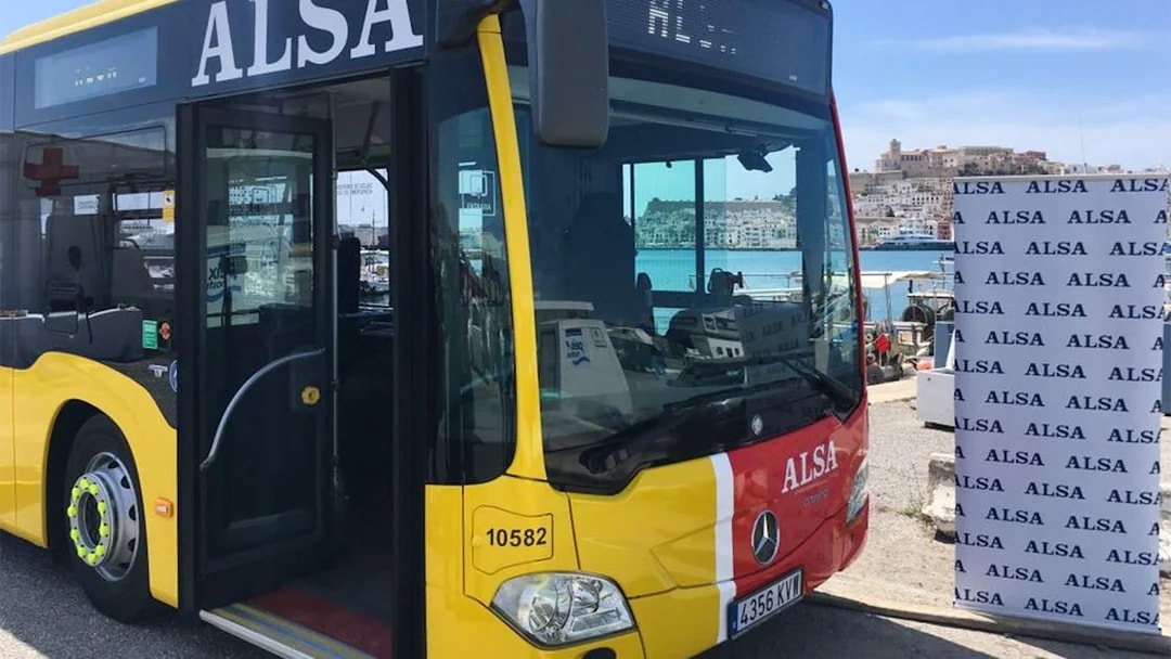 Yellow ALSA bus at Ibiza port with city view in background.