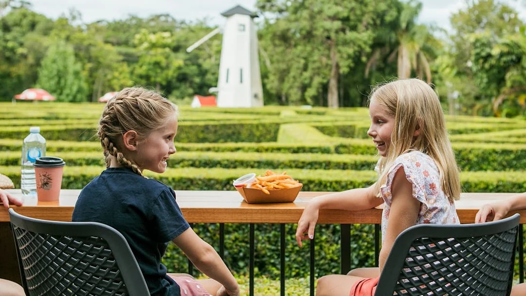 Children enjoying snacks at a table with a hedge maze and windmill in Amaze World.