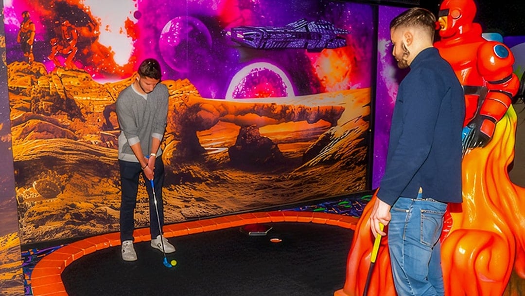 Guests playing mini golf under blacklight with a sci-fi themed backdrop.