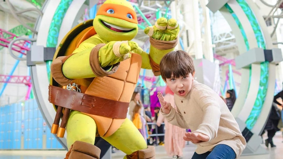 Child posing with Teenage Mutant Ninja Turtle character at an indoor amusement park.